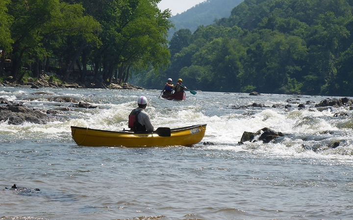 In the background, two students paddle a canoe toward whitewater. In the foreground, another person, likely an instructor, watches them from a canoe.